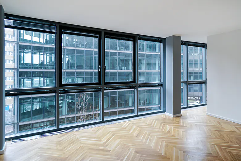 Bright, empty room with herringbone wood floors and large windows. Outside, a tall office building is visible.