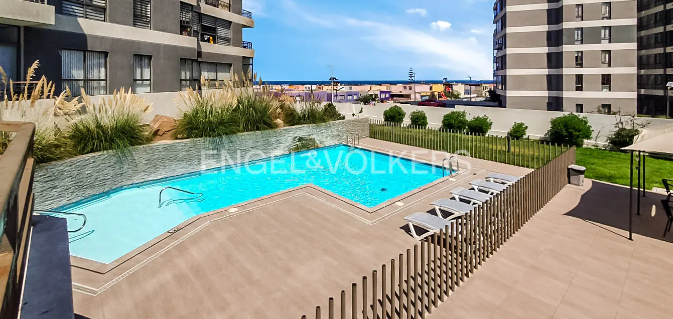 Elevated view of a blue pool with lounge chairs, a green lawn, and buildings under a blue sky.