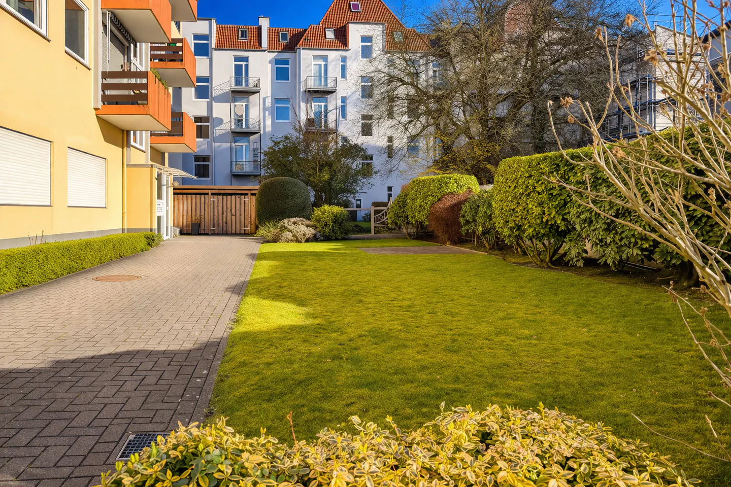 Apartment complex with a green lawn, trimmed hedges, and a brick walkway. The buildings are yellow and white with orange balconies.