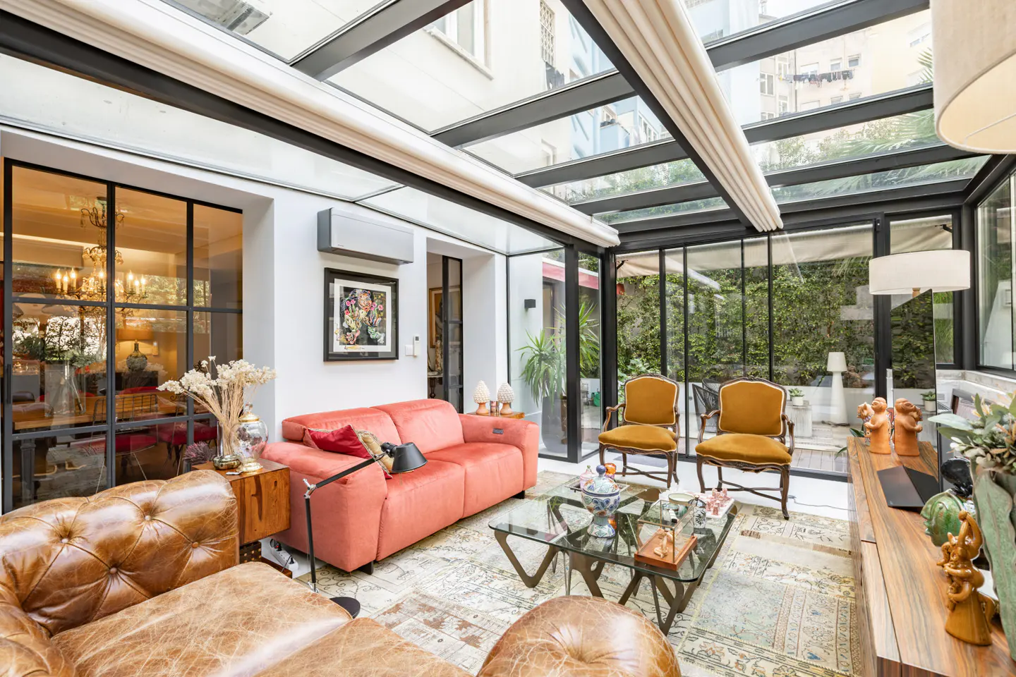 Living room with glass ceiling, coral sofa, and leather chairs. Natural light floods the space with a view of greenery outside.