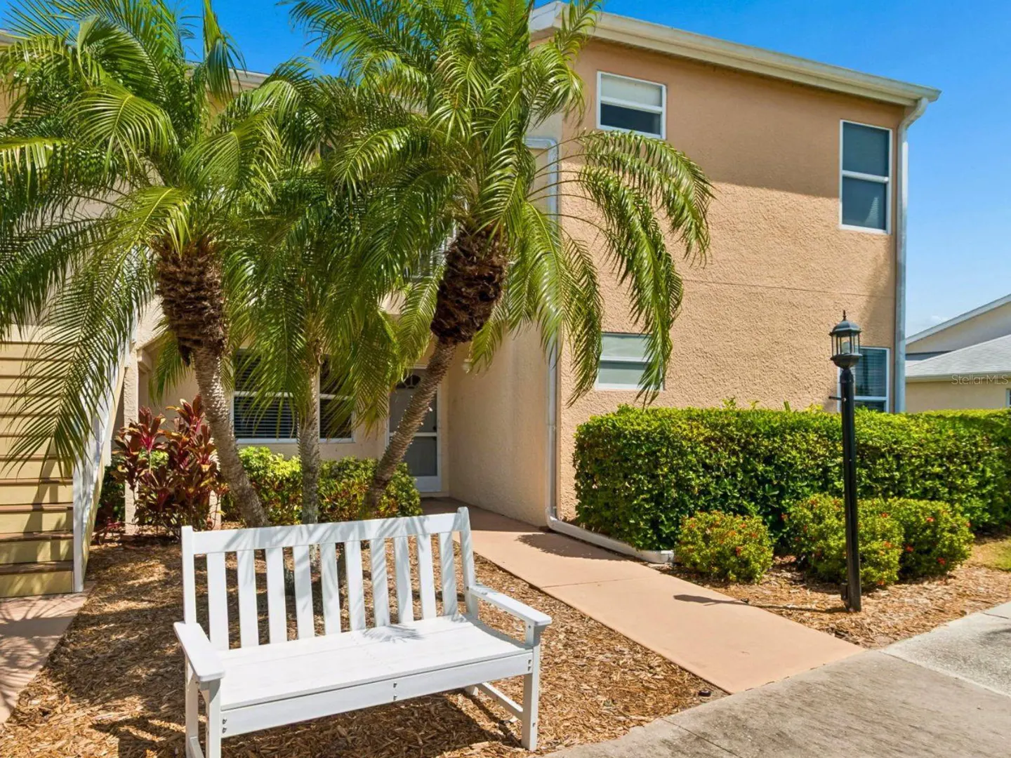 Exterior of a tan two-story building with palm trees, a white bench, and green bushes.