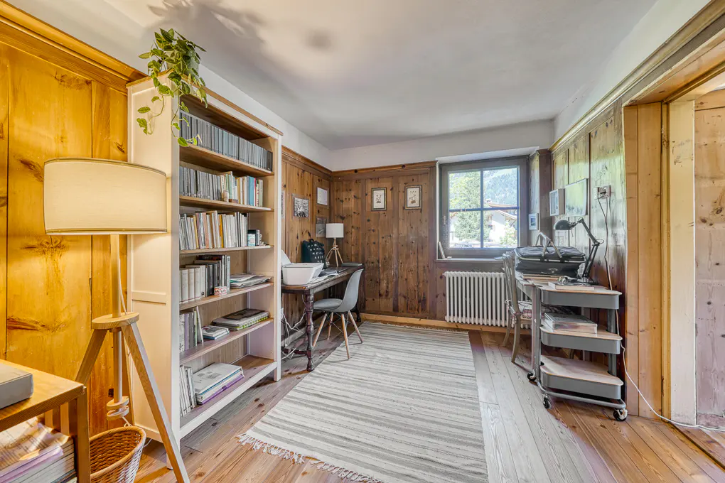 A home office with wood paneling, a white bookcase filled with books, and two desks. A striped rug covers the wood floor.