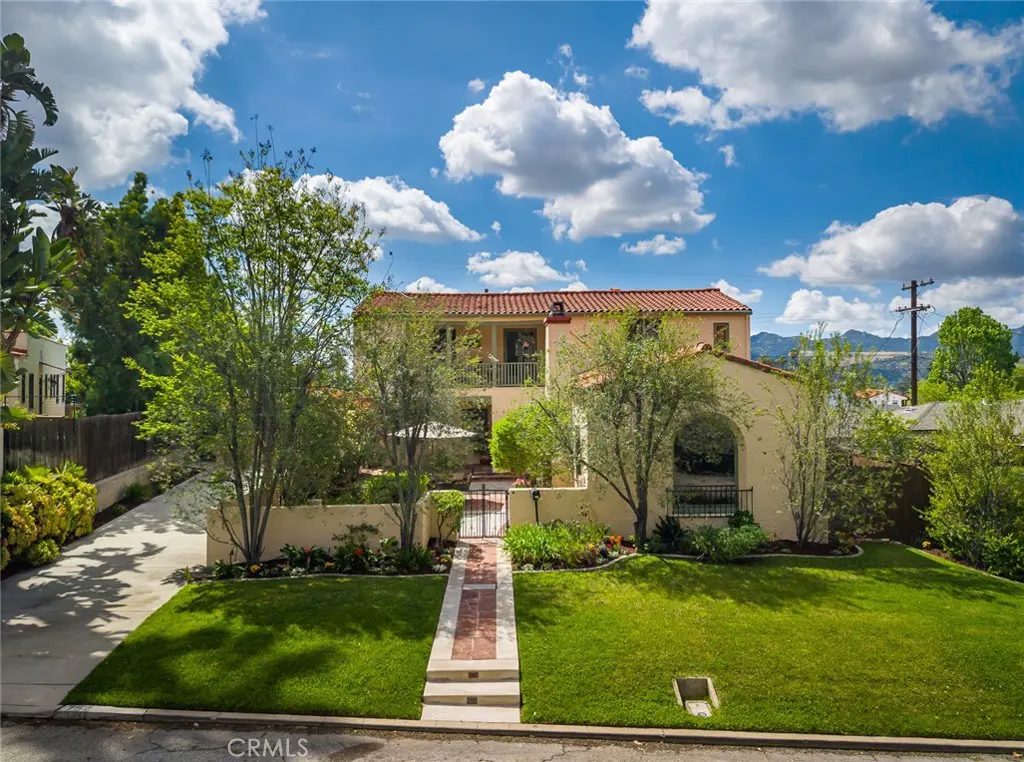 Two-story beige house with a red tile roof, green lawn, trees, and a brick walkway under a blue sky with white clouds.