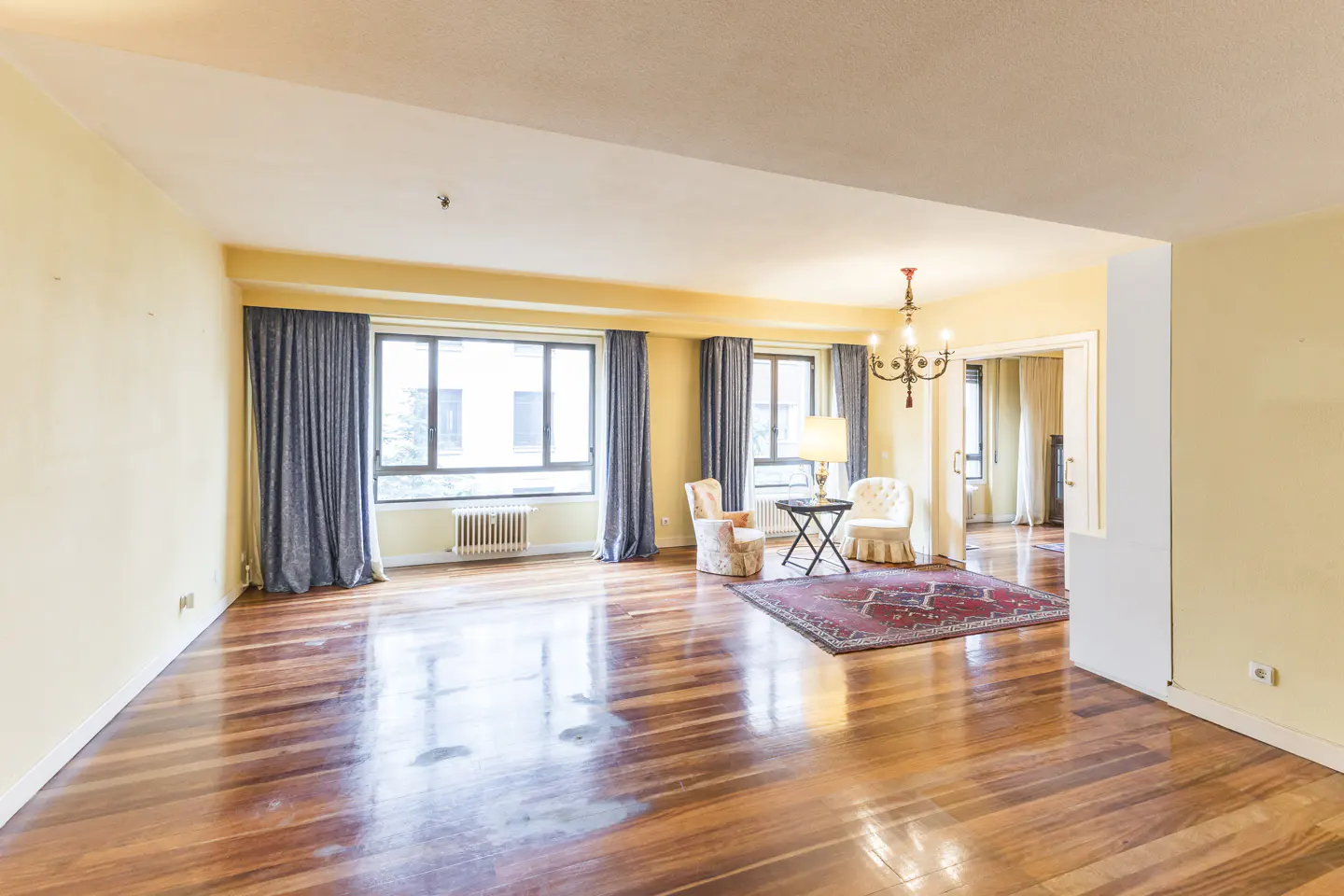 A bright living room with hardwood floors, yellow walls, and blue curtains. Two chairs and a table sit on a red rug near large windows.
