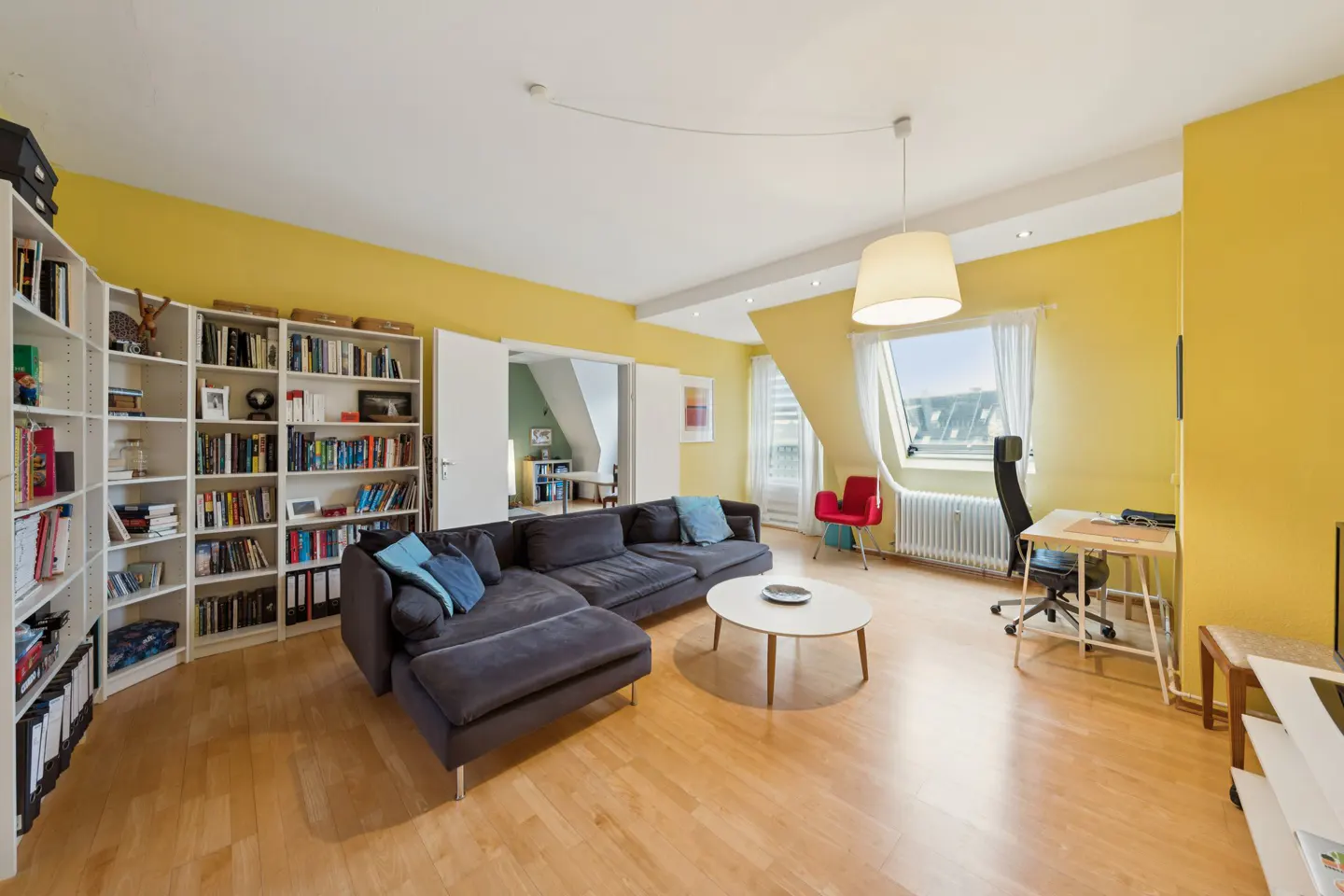 Bright living room with yellow walls, wood floors, and a large bookcase. A gray sectional sofa faces a round coffee table. A desk sits near a window.