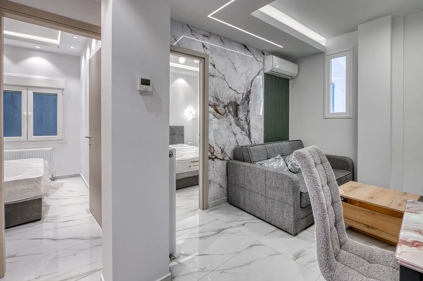 A modern apartment interior with marble floors, a gray sofa, and a view into a bedroom.