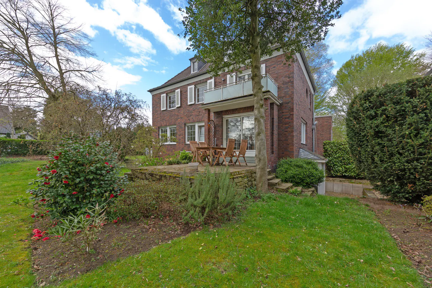 Brick house with white trim, a balcony, and a patio with a table and chairs, surrounded by green grass and trees.