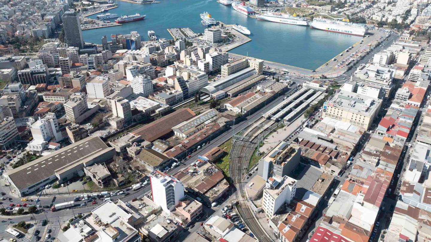 Aerial view of Piraeus port in Greece, showing city buildings, train tracks, and large ferries docked in the blue harbor.