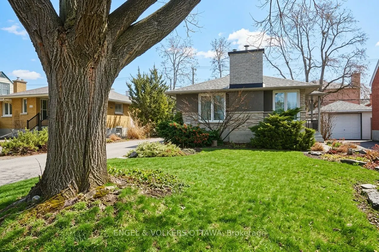Exterior view of a single-story house with a stone chimney, green lawn, and a large tree in the foreground.