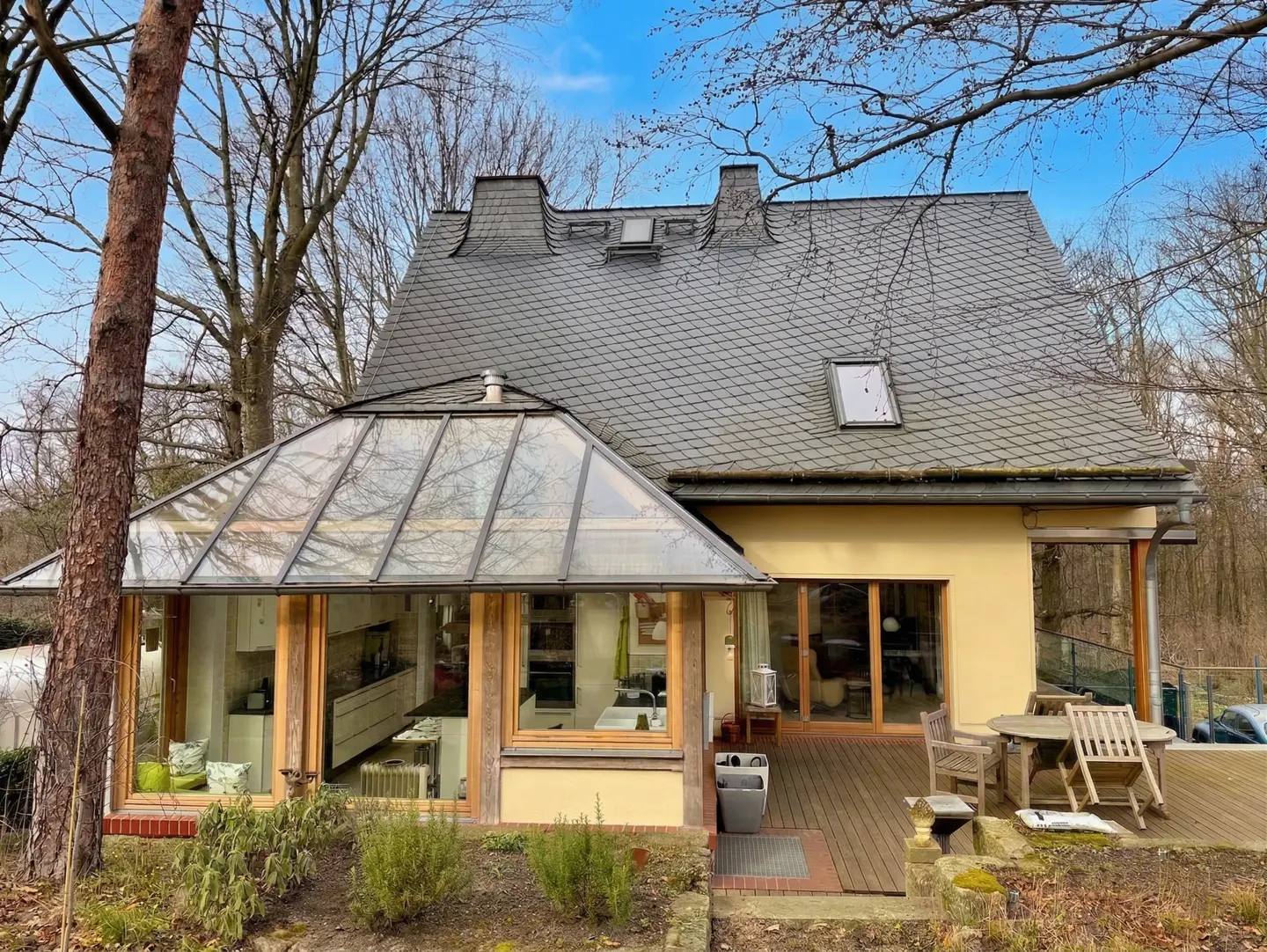 Exterior view of a yellow house with a glass conservatory, gray roof, and wooden deck with outdoor furniture.