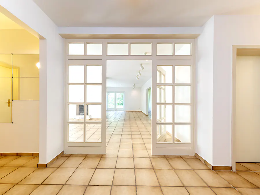 Interior view of a home with tan tile flooring, white walls, and a white framed doorway leading to a bright living room.