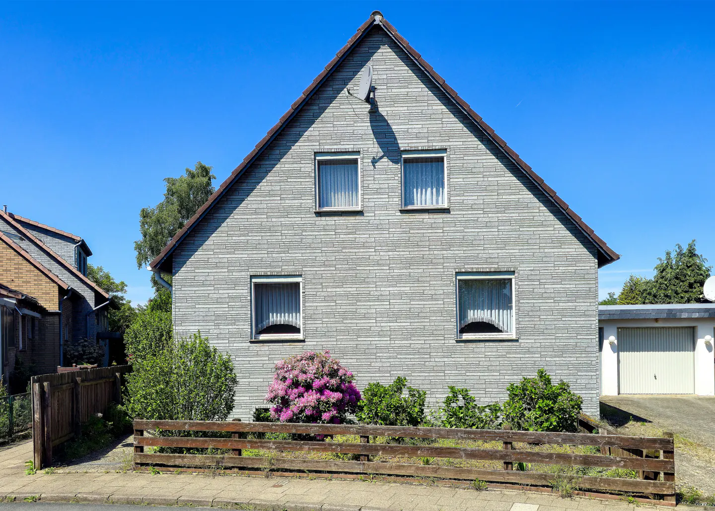 A two-story gray house with a brown roof and white-framed windows, under a clear blue sky. A wooden fence and pink flowers are in front.