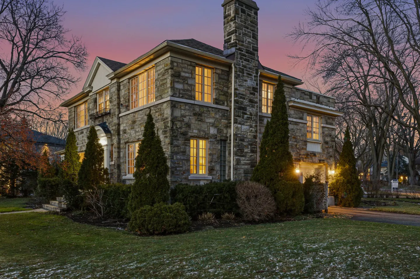 Exterior view of a large stone house at dusk with lit windows, green lawn, and bare trees.