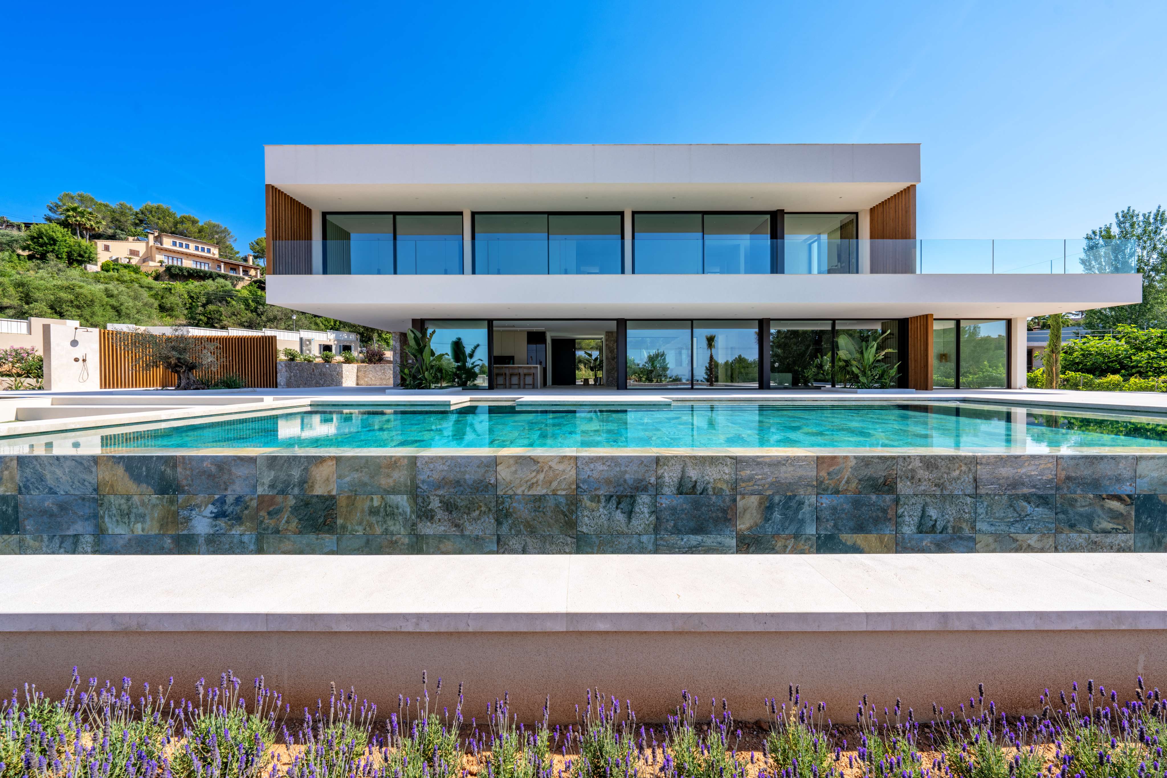 Modern two-story villa with a turquoise infinity pool, stone-tiled wall, and lavender plants in the foreground under blue sky. [AI-generated text]