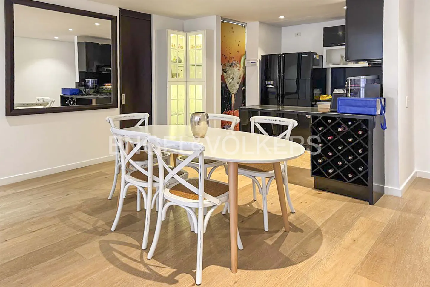 Modern dining area with a white oval table, four white chairs, a large wall mirror, and a black kitchen island with wine rack.
