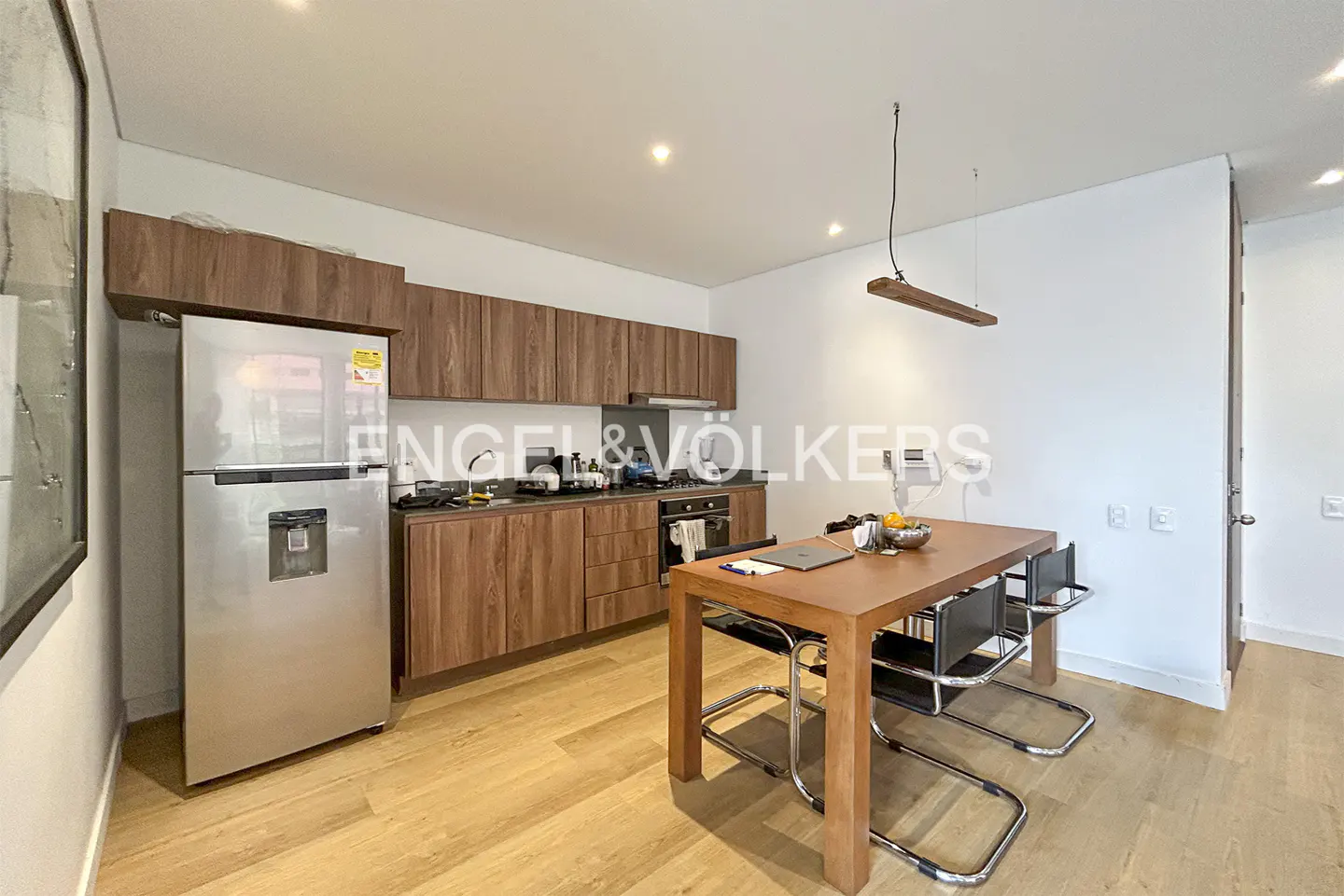 A modern kitchen with wood cabinets, stainless steel refrigerator, and a wood dining table with four chairs.