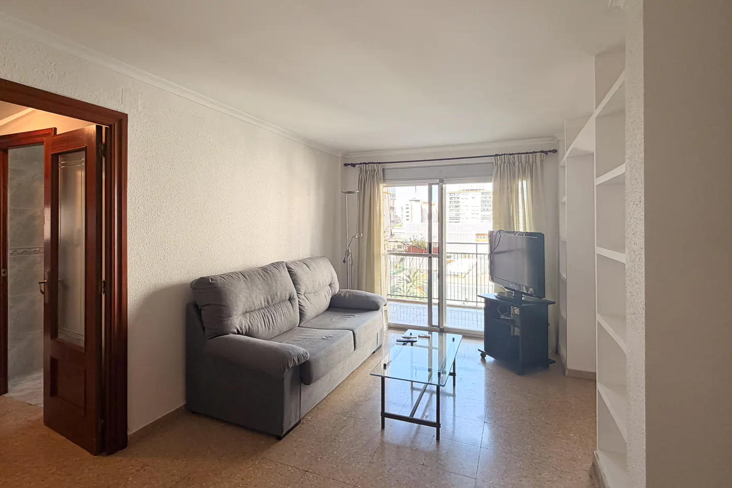 Living room with a gray sofa, glass table, TV, and balcony with city views. A wooden door and white shelves are also visible.