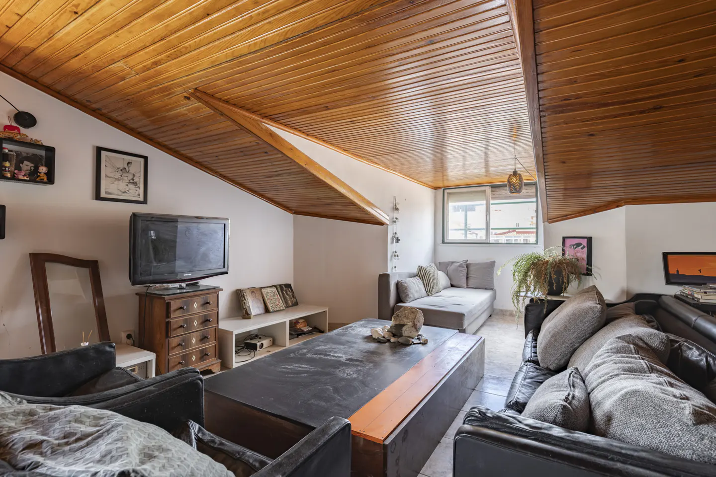 Attic living room with wood-paneled ceiling, black leather sofas, and a large coffee table. A TV sits on a wooden dresser.