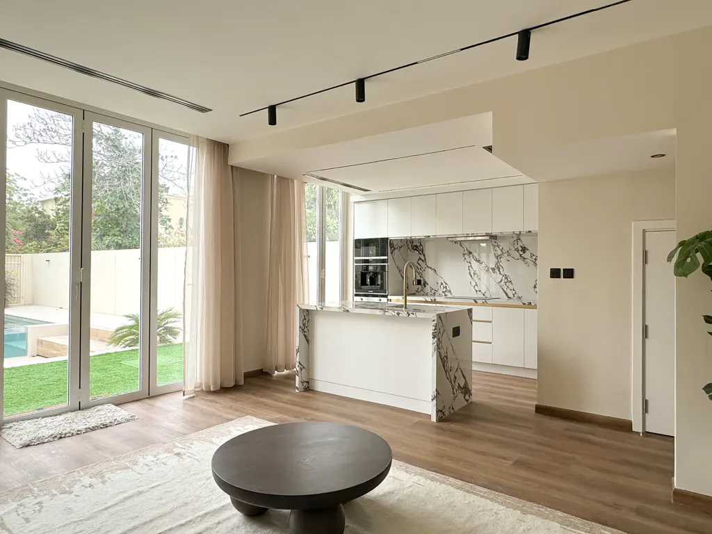 Bright, modern kitchen and living area with white cabinets, marble countertops, and a large window overlooking a pool.