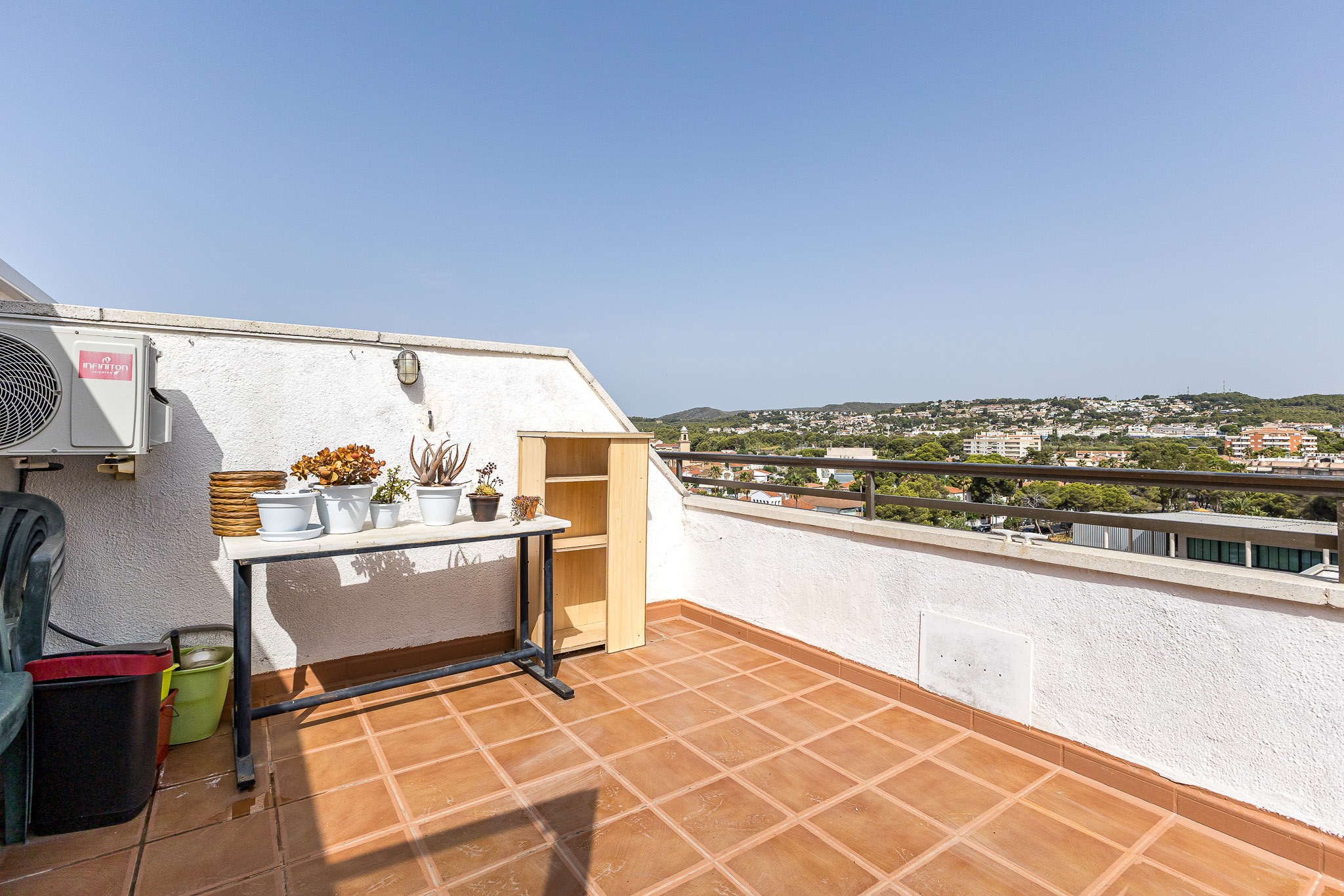 Sunny balcony with terracotta tiles, a table holding potted plants, a small wooden shelf, and a view of a distant town.