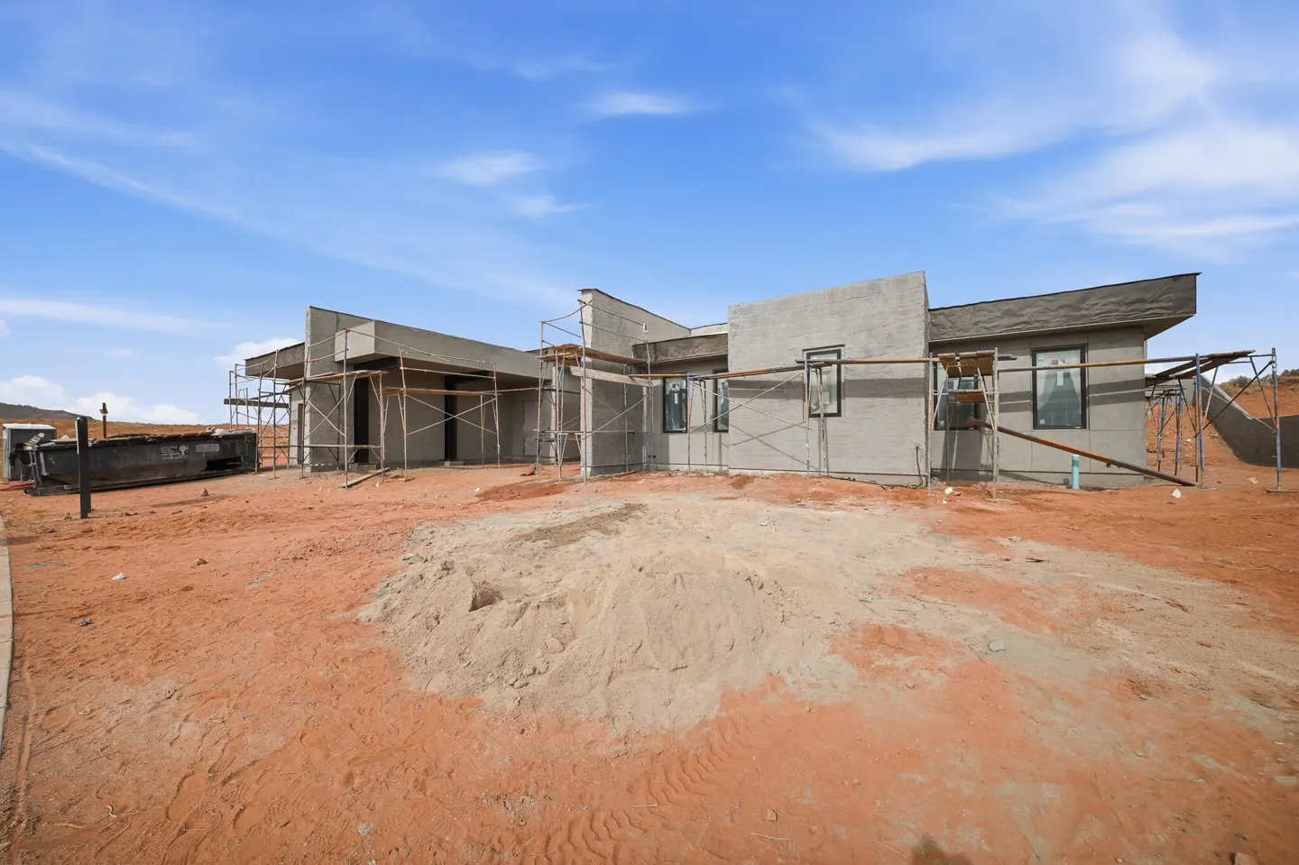 Exterior view of a modern, gray stucco house under construction with scaffolding and a red dirt yard under a blue sky.