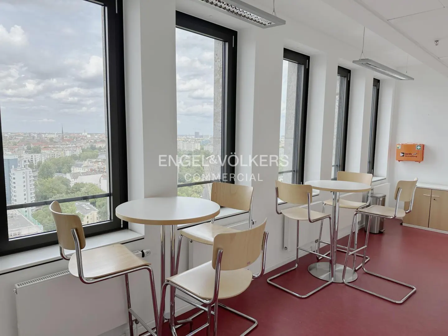 Bright office space with tables and chairs by large windows overlooking a city. Red floor, white walls, and Engel & Völkers logo visible.