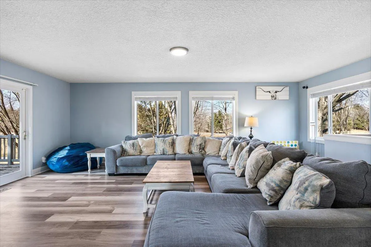 Living room with a large gray sectional sofa, wood floors, and light blue walls. Windows overlook a wooded area. A blue beanbag chair sits near a sliding glass door.