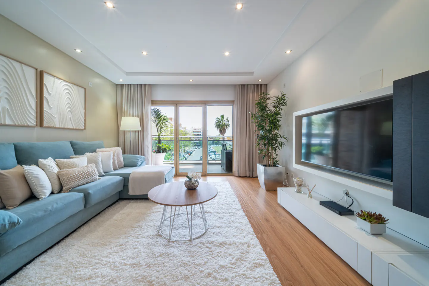 Bright living room with a blue sectional sofa, white rug, and a round coffee table. A large TV is mounted on the wall, and a balcony is visible through sliding glass doors.