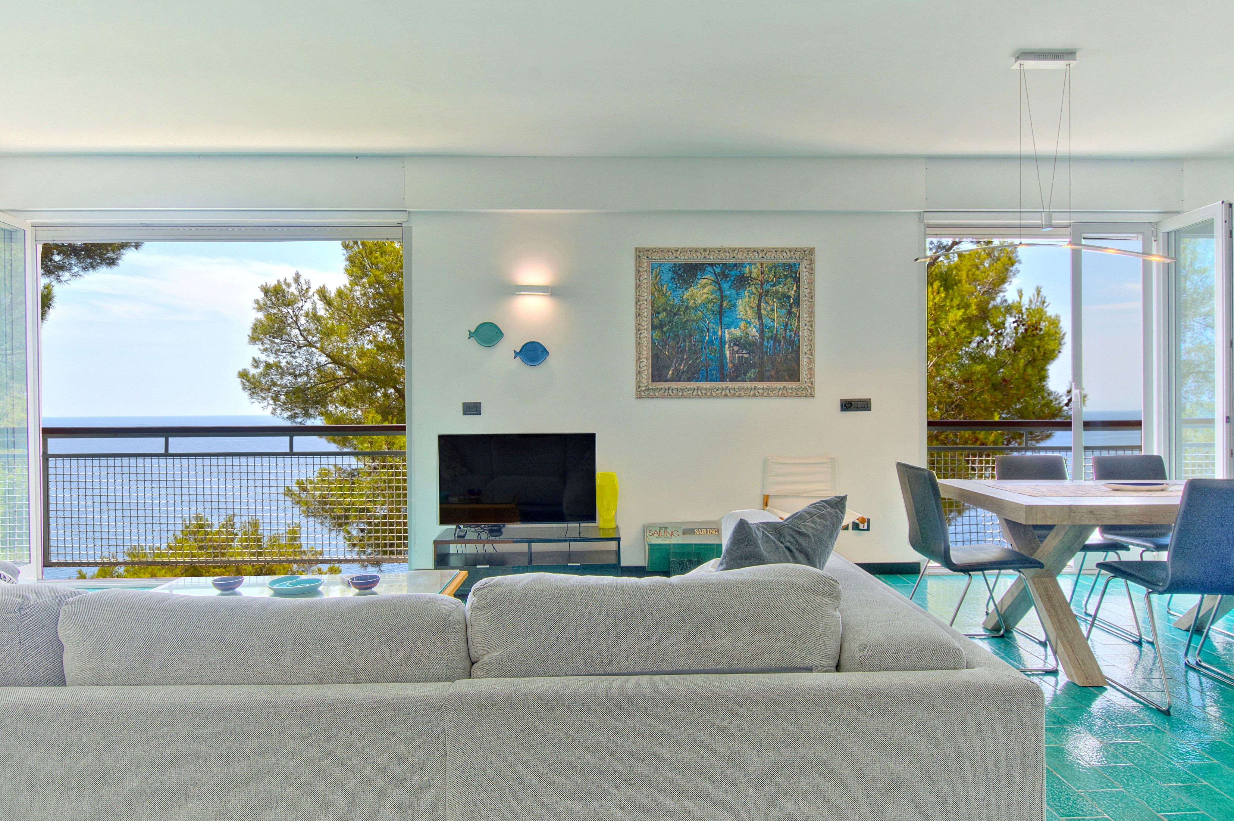 Bright living room with a white sofa, TV, dining area, and large windows overlooking the sea and pine trees.