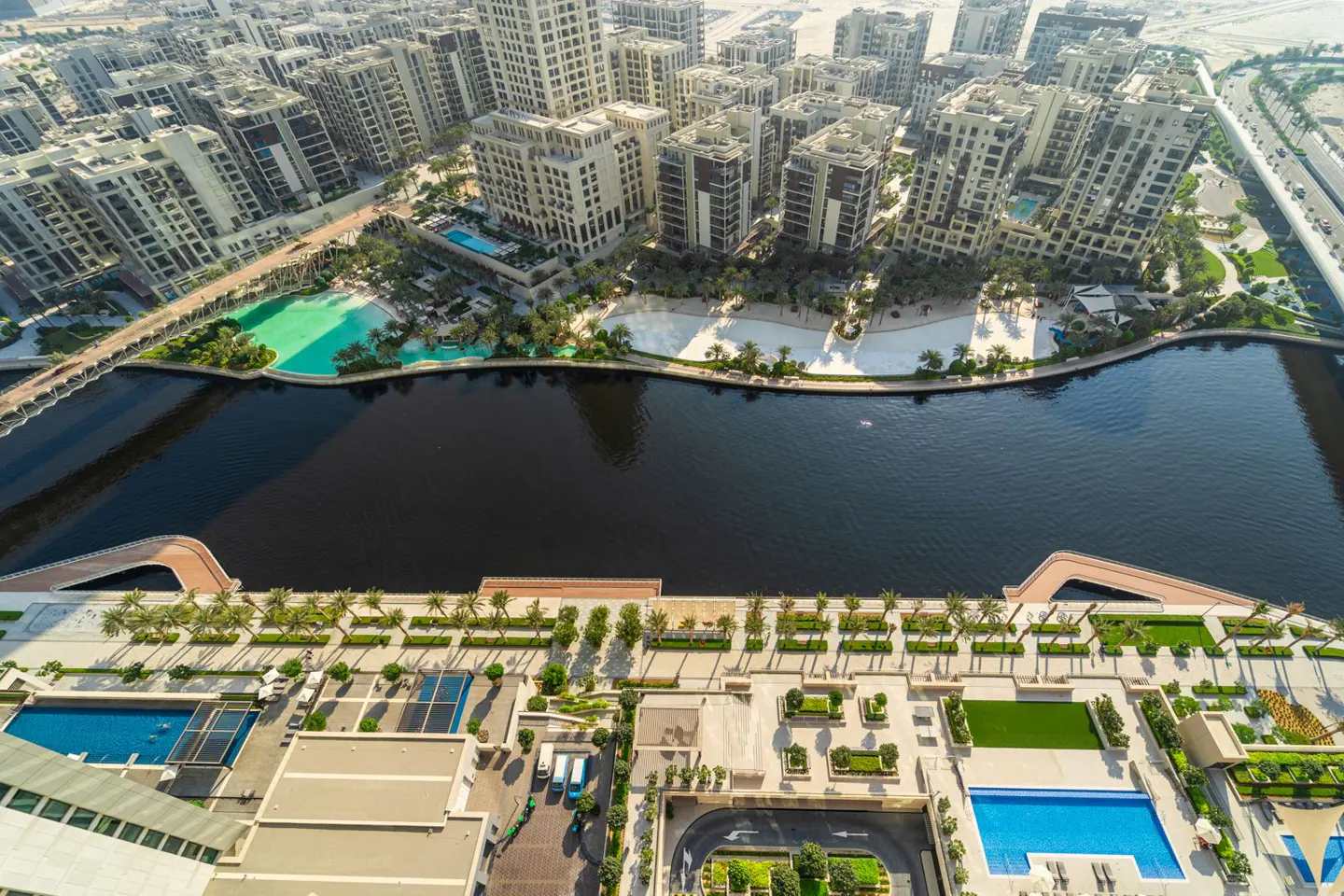 Aerial view of a luxury residential area with beige buildings, a dark river, and turquoise pools. Palm trees and manicured lawns add to the upscale ambiance.