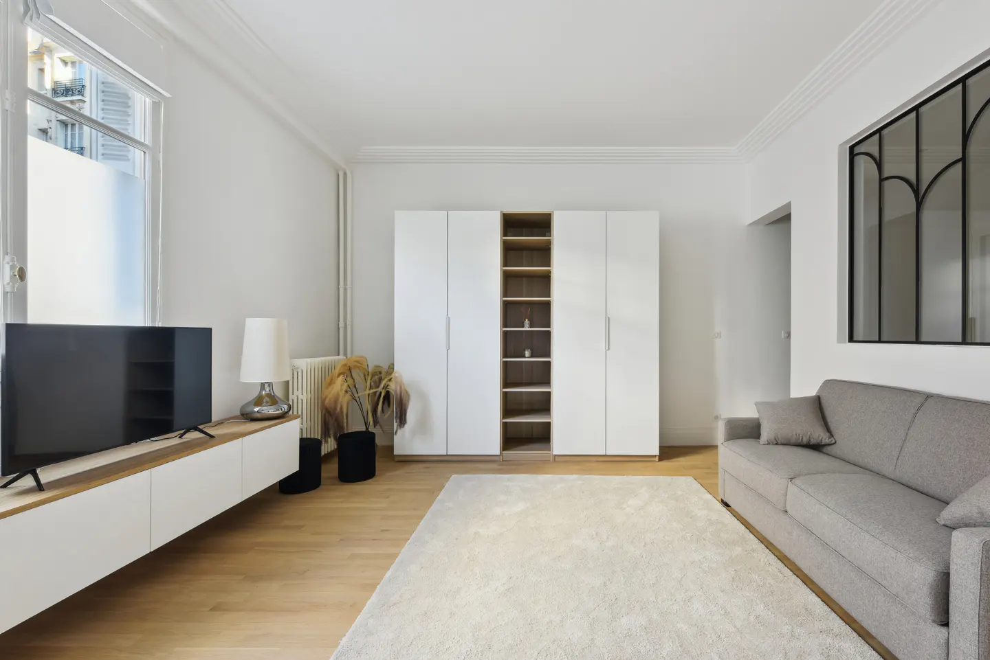 Bright living room with a gray sofa, white cabinets, a TV on a console, and a large window.