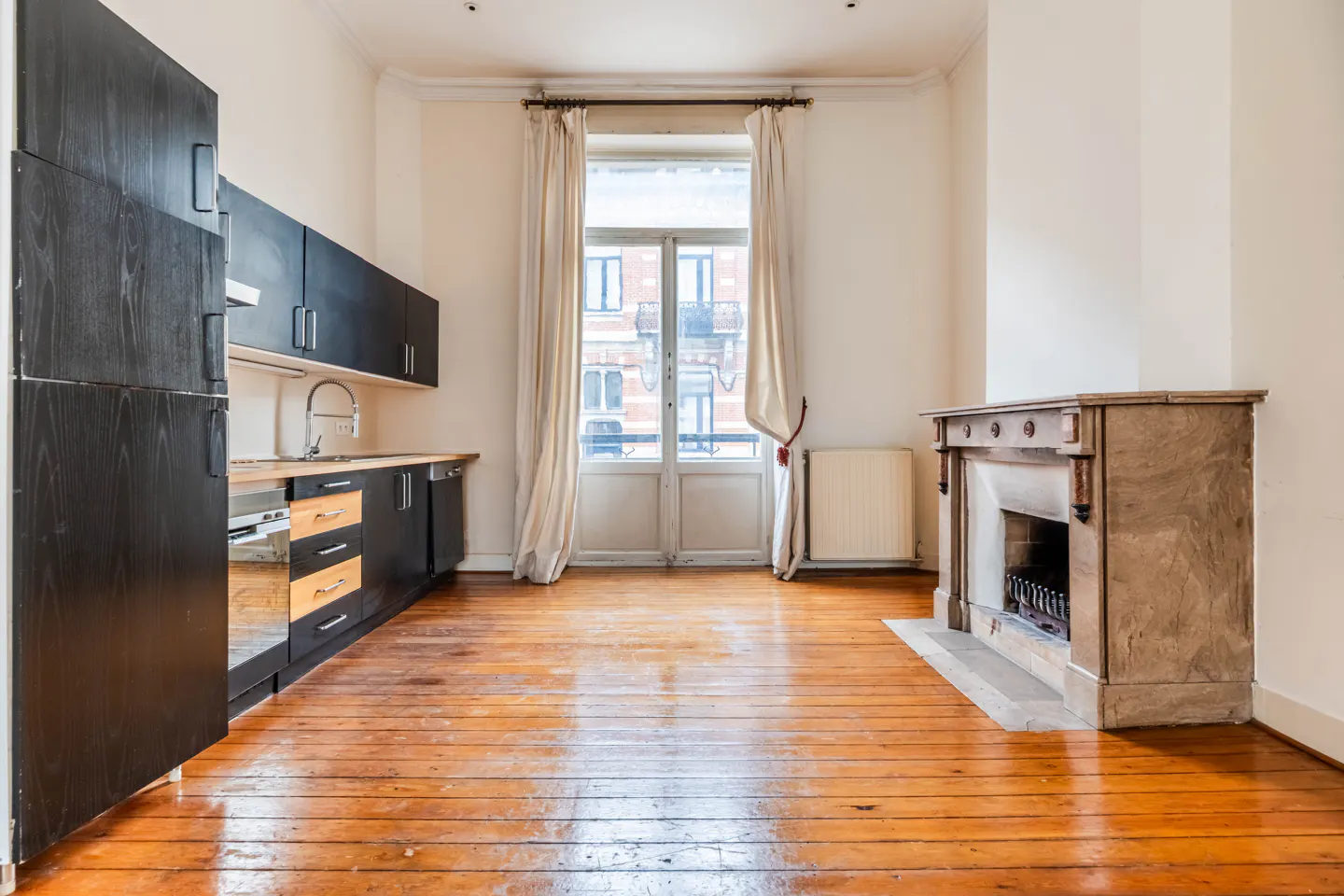 A bright room with wood floors, a black kitchen, a stone fireplace, and a window with white curtains.