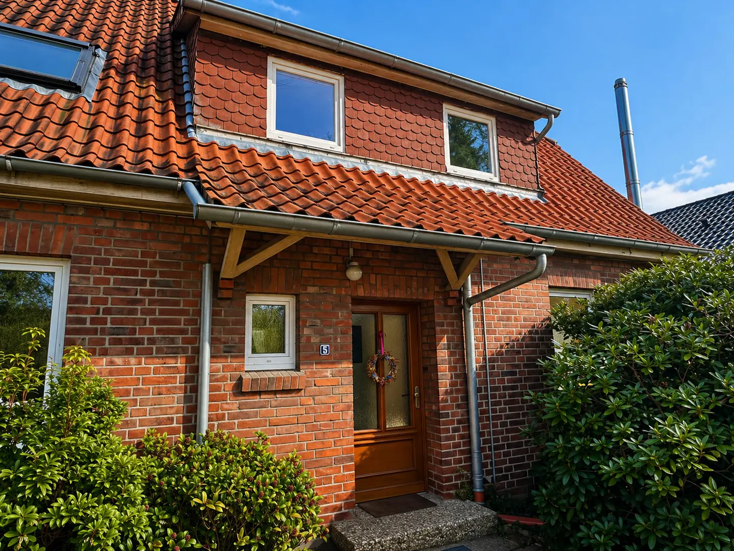 Red brick house with a brown door, red tile roof, and green bushes. A wreath hangs on the door.