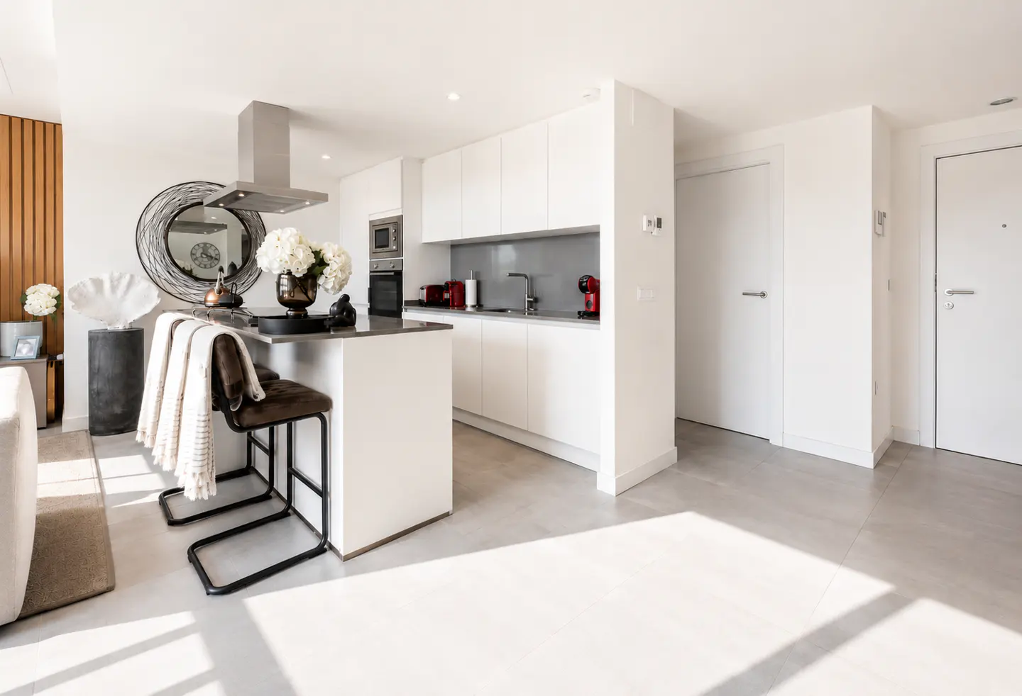 Bright, modern kitchen with white cabinets, island with stools, and stainless steel appliances. Sunlight streams across the gray floor.