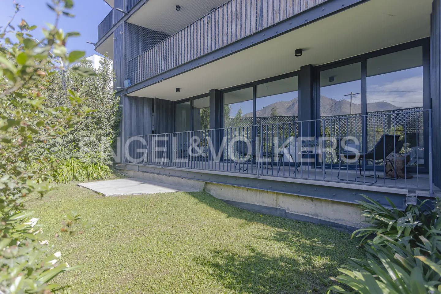 Exterior view of a modern apartment building with a balcony overlooking a green lawn and distant mountains.