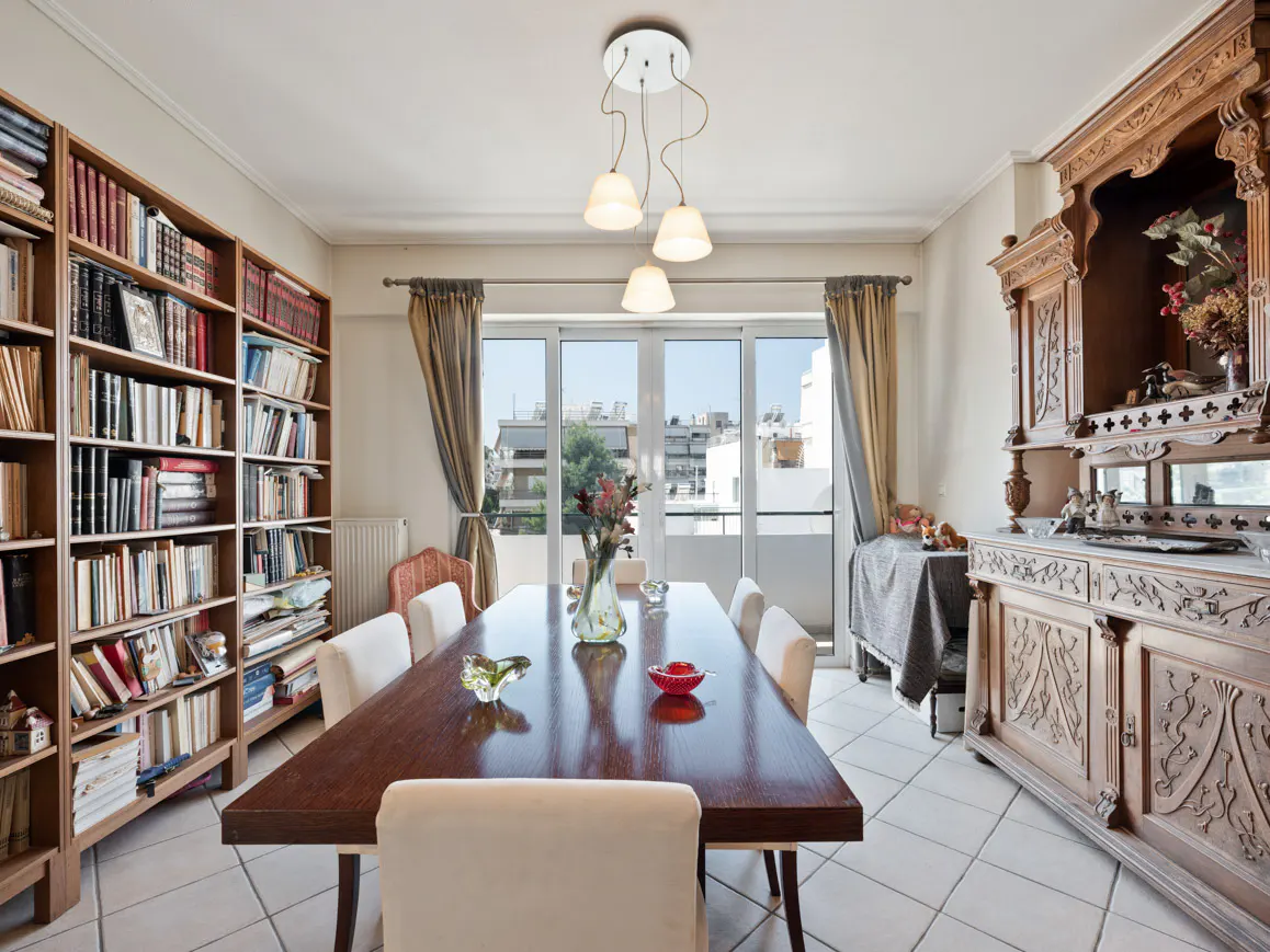 A dining room with a long wooden table, white chairs, a bookshelf, and a carved wooden cabinet.