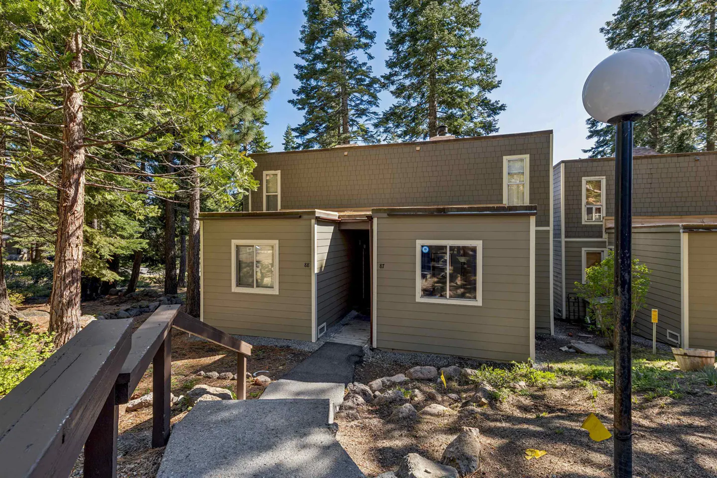 Exterior view of a two-story condo with olive green siding and a brown shingled roof, surrounded by trees. A stone walkway leads to the entrance.