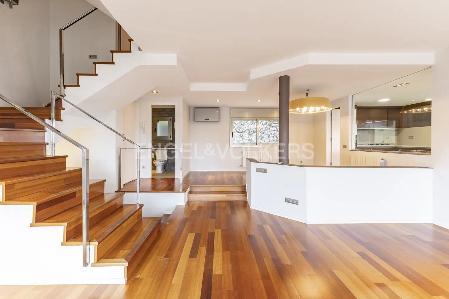 Open-concept home interior with wood floors, white walls, and a staircase with stainless steel railings. A kitchen is visible in the background.