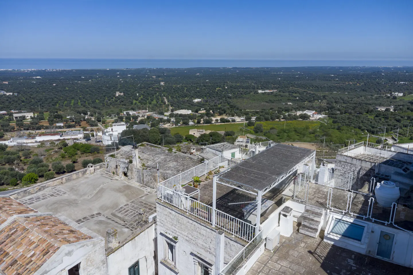 Aerial view of a white stone building with a rooftop terrace, overlooking green fields and the ocean under a blue sky.