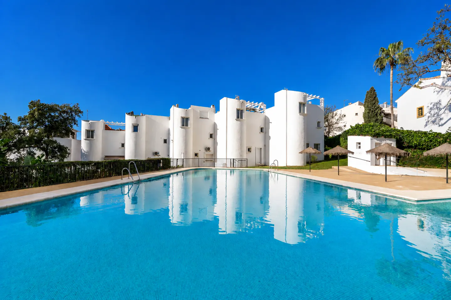 A turquoise pool reflects white buildings under a clear blue sky. Palm trees and sun umbrellas line the pool's edge.