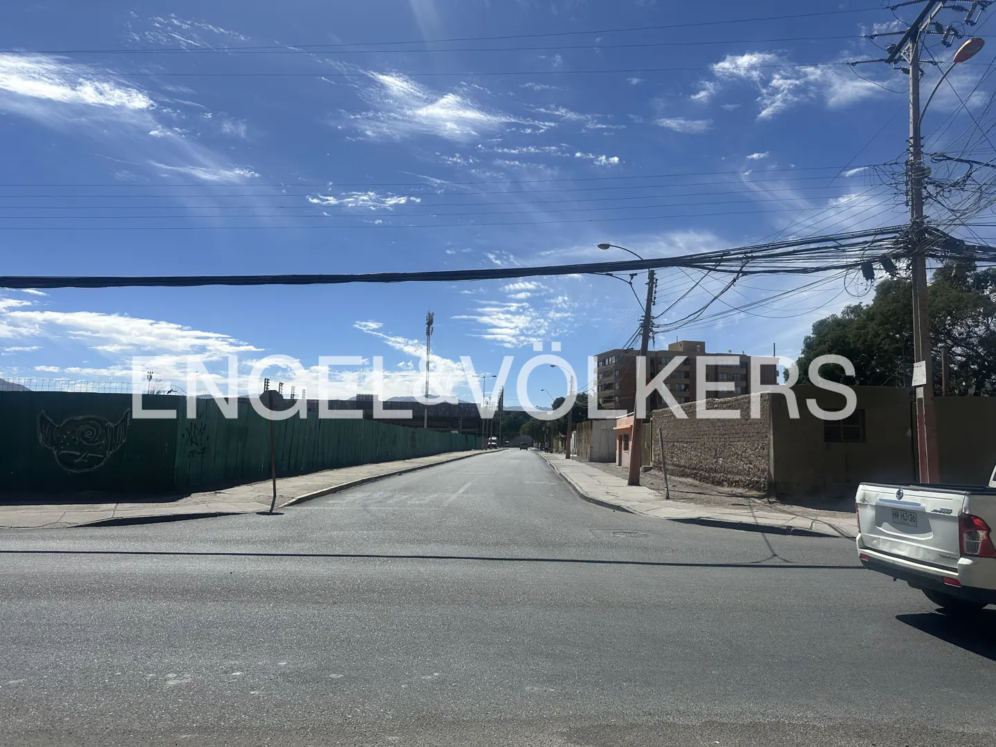 Street view with a silver pickup truck on the right, a green fence on the left, and a blue sky with clouds above.