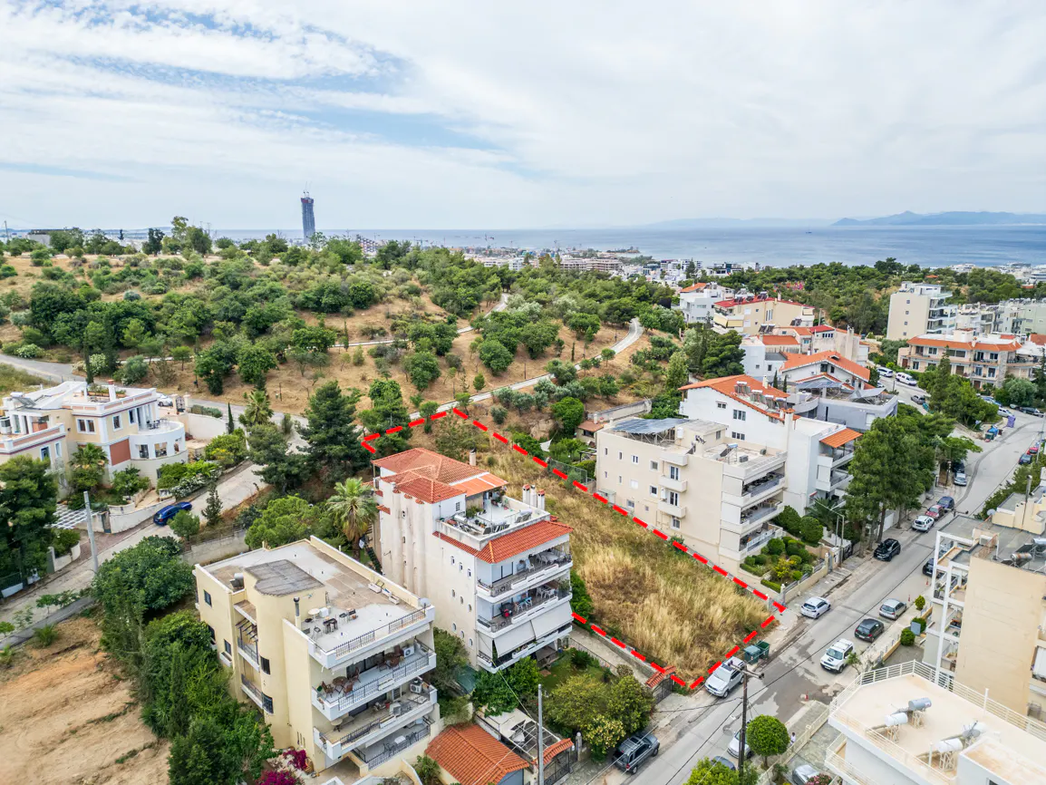Aerial view of a vacant residential lot outlined in red, situated between apartment buildings near a coastal hillside.