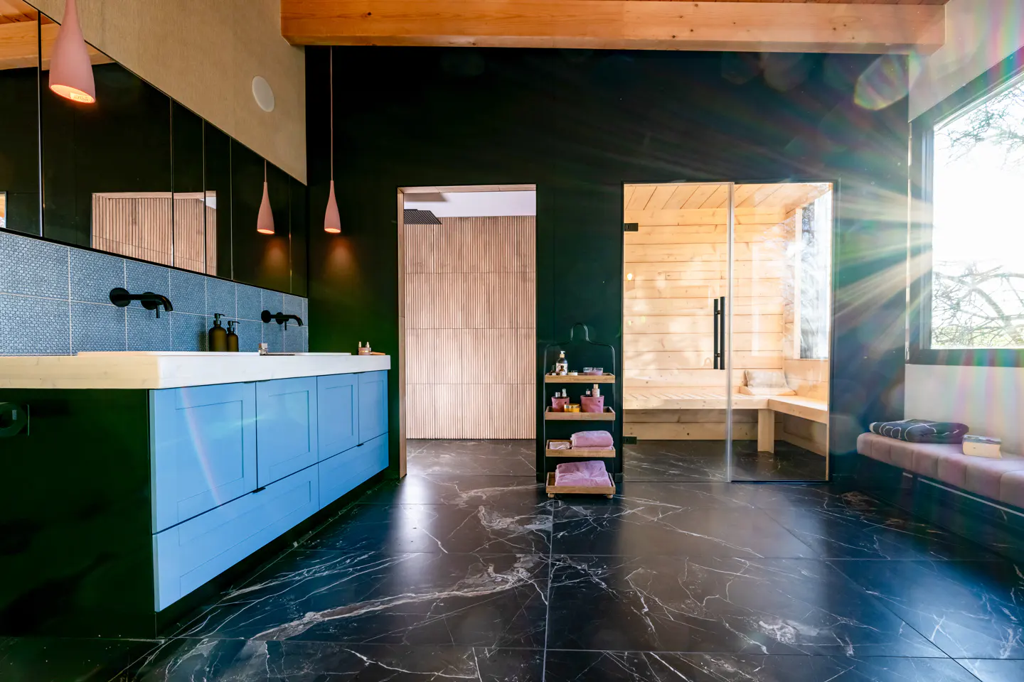 Modern bathroom with blue vanity, black marble floors, and a glass-enclosed wooden sauna under a natural wood ceiling beam.