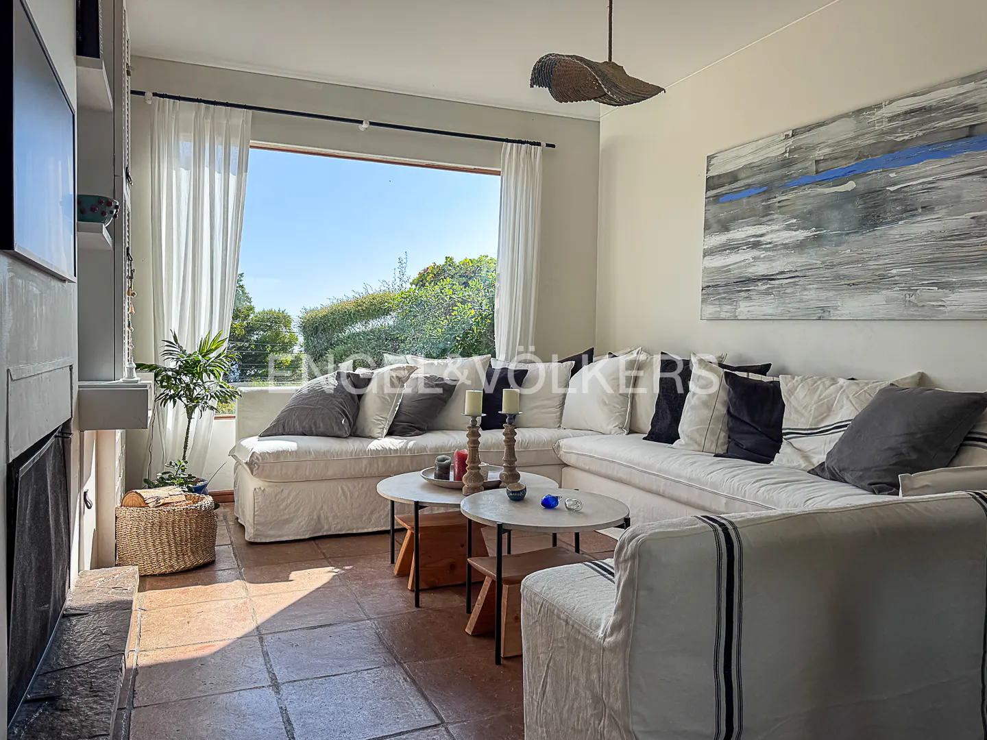 Living room with white sectional sofa, pillows, and round tables. Large window with view of greenery. Abstract art on the wall.