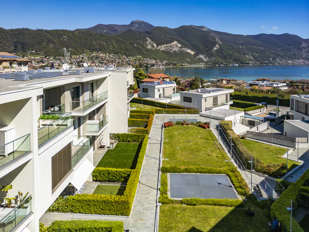 Aerial view of modern white apartments with glass balconies, green lawns, and a lake and mountains in the background.