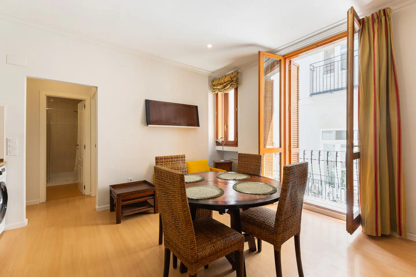 Bright dining area with a round wooden table, four wicker chairs, and open doors to a balcony with a black railing.