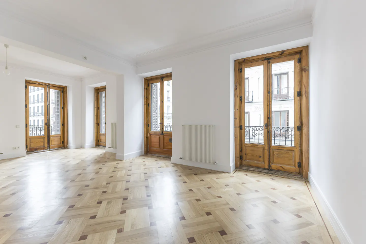Bright, empty room with parquet floors, white walls, and three sets of wooden doors leading to balconies. A white radiator is visible.