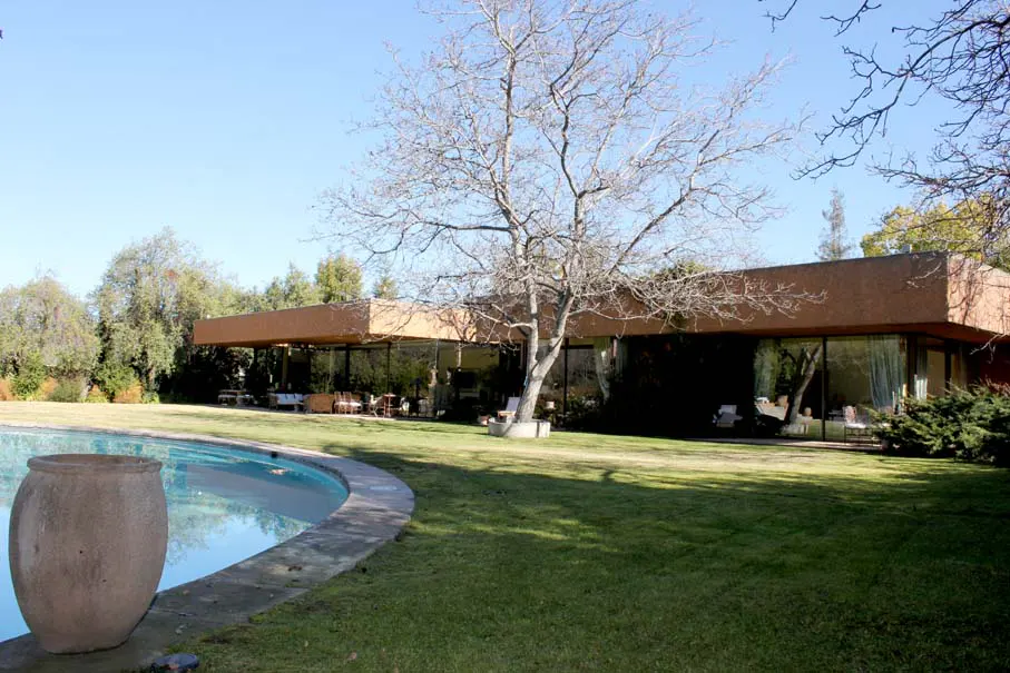 Modern house with a pool. The house has a brown exterior and large glass windows. A bare tree stands in front of the house.