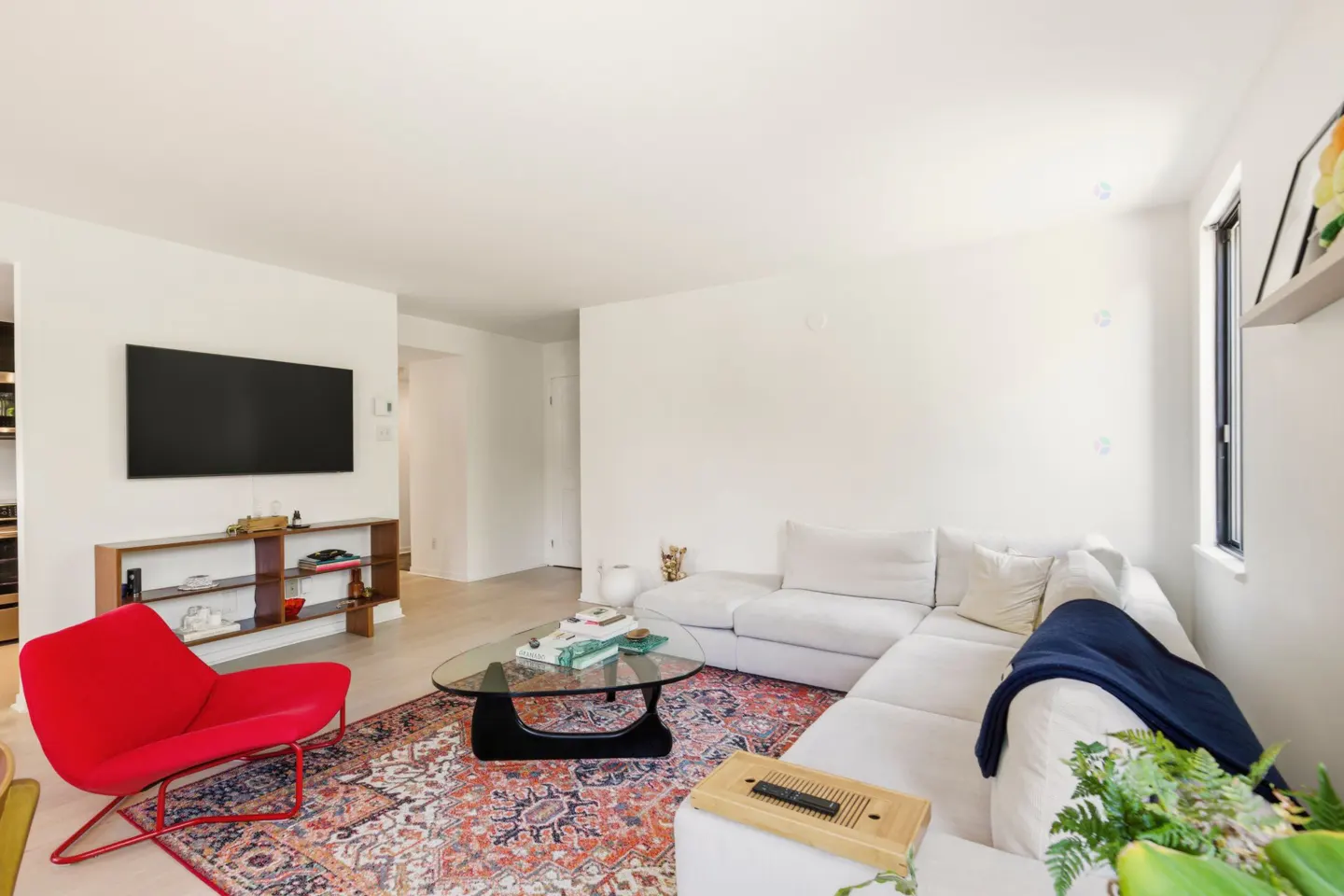 Bright living room with white walls, a large white sectional sofa, a red chair, and a patterned rug. A TV hangs above a wooden shelf.