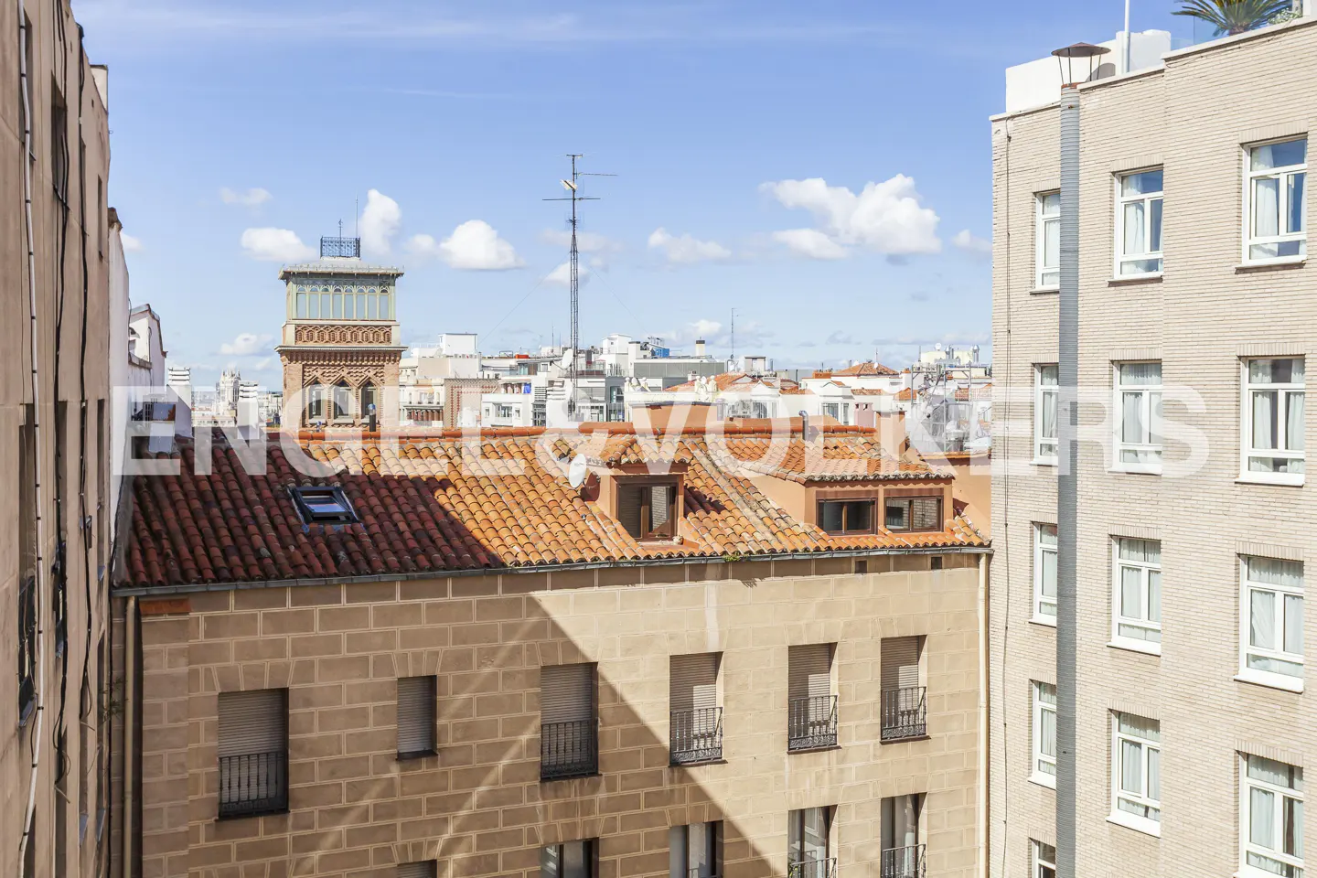 Cityscape view of Madrid, Spain, featuring buildings with red tile roofs and a brick tower under a blue sky with scattered clouds.